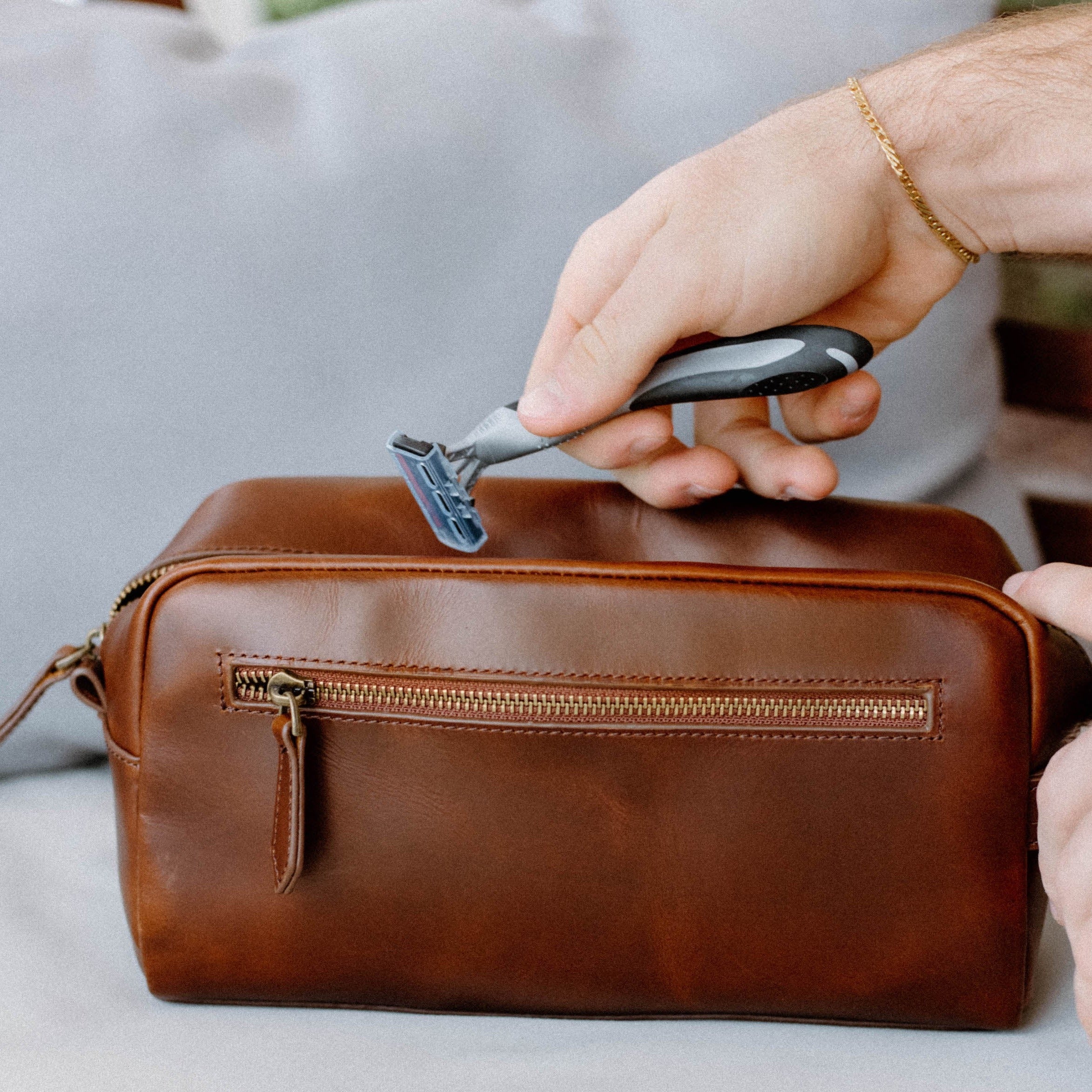 A person holding a razor above a brown leather Dopp Kit bag.