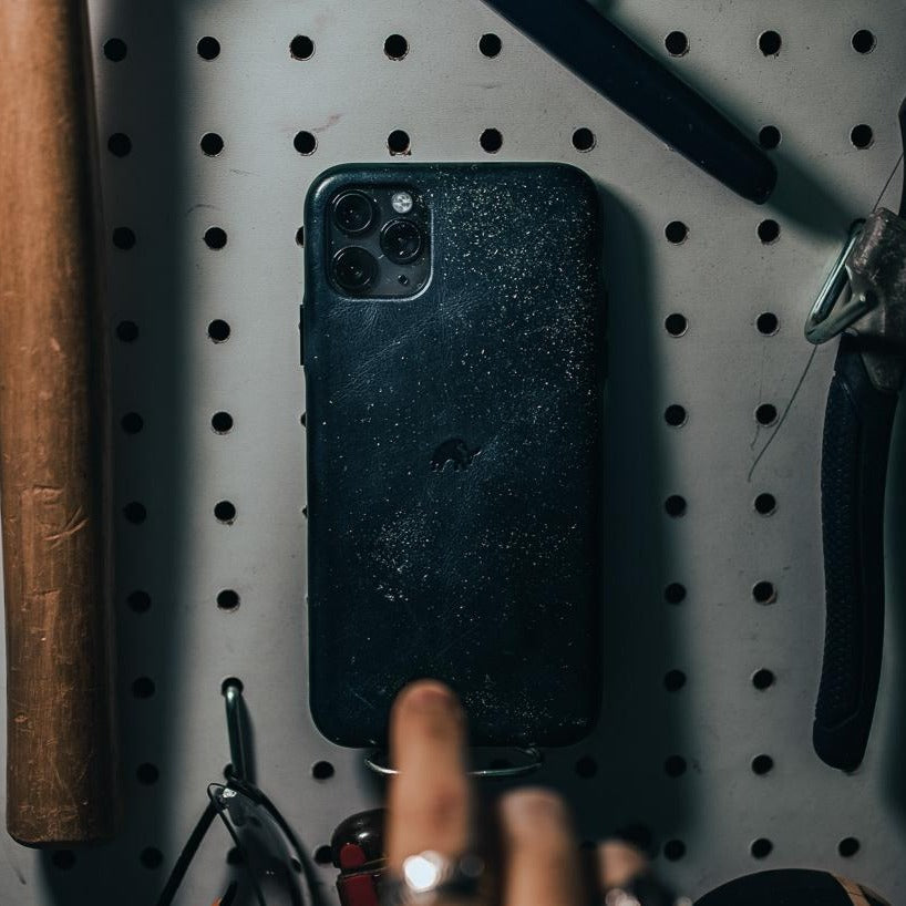 Worn dark ocean-colored iPhone case hanging on a pegboard hook with a blurred hand reaching; tools visible nearby.