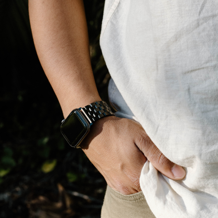 Left wrist wearing a black metal link Apple Watch band; hand in pocket, casual white shirt visible.