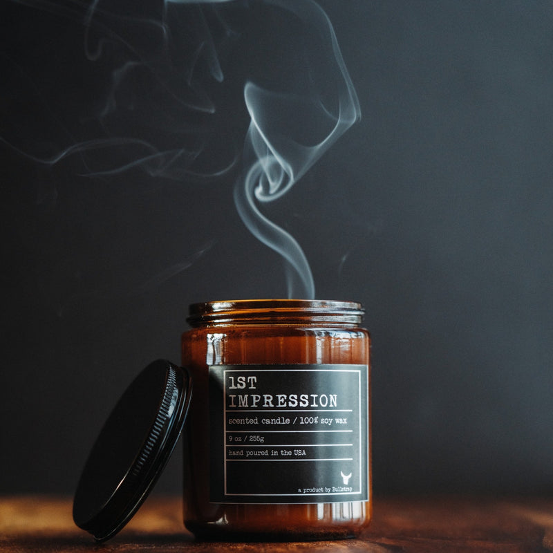 Front view of 1st Impression 9 oz candle in amber jar showing label 1ST IMPRESSION and smoke against a dark background