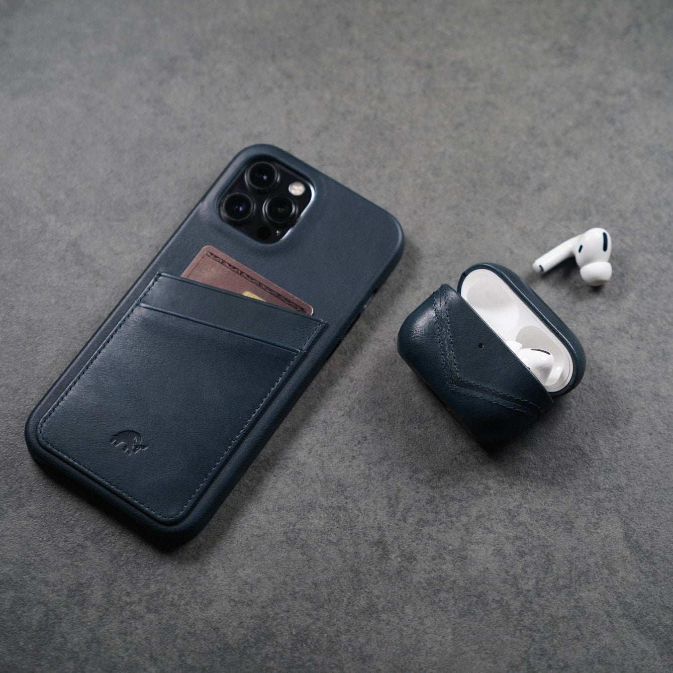 Dark blue leather phone case with card slot beside an open dark blue leather AirPods case holding white AirPods.