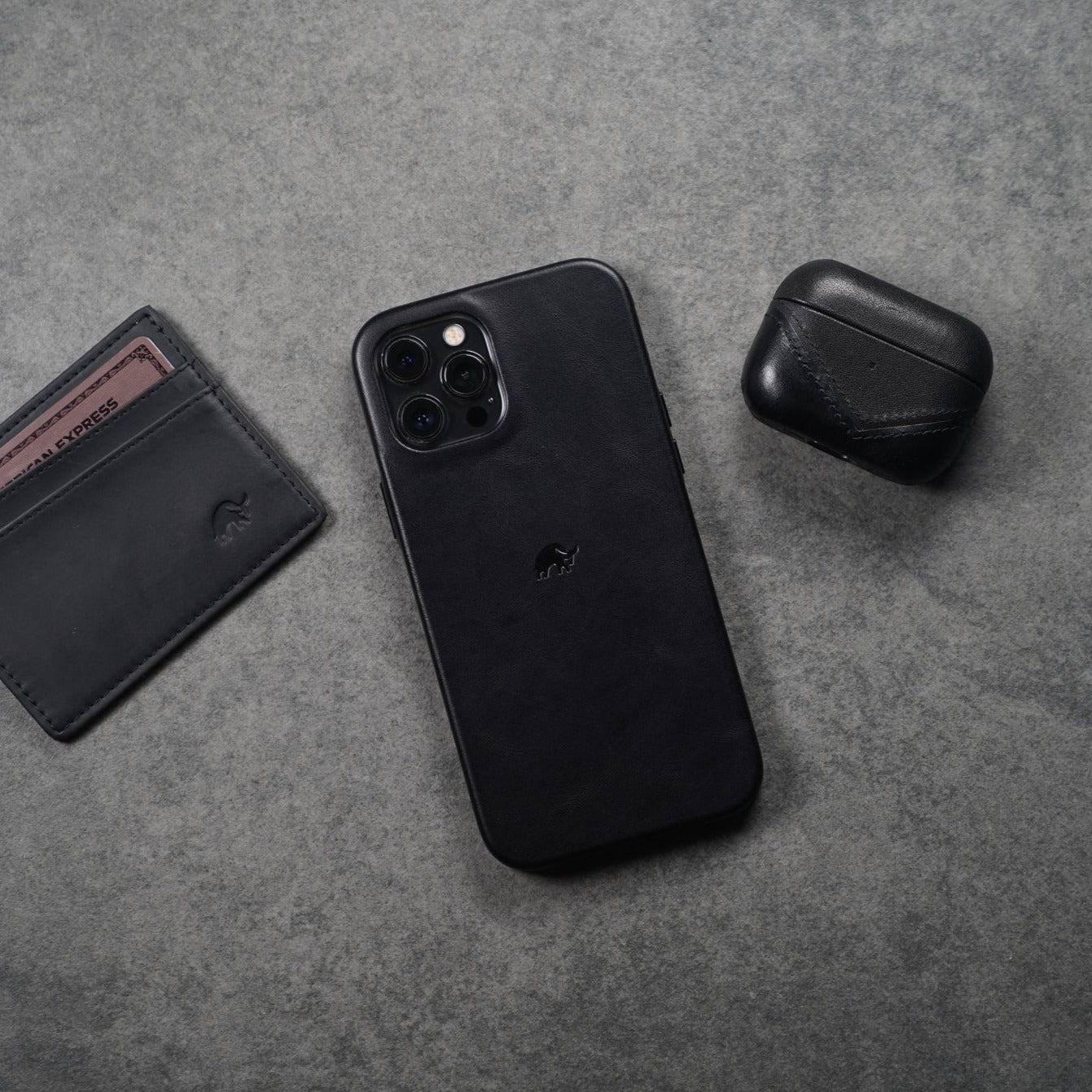 Black leather AirPods case next to a phone and wallet on a gray surface.