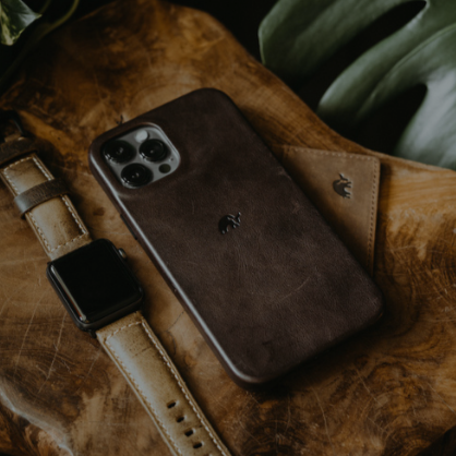 Brown leather iPhone case on a wooden tray with a matching leather card holder and tan watch band.