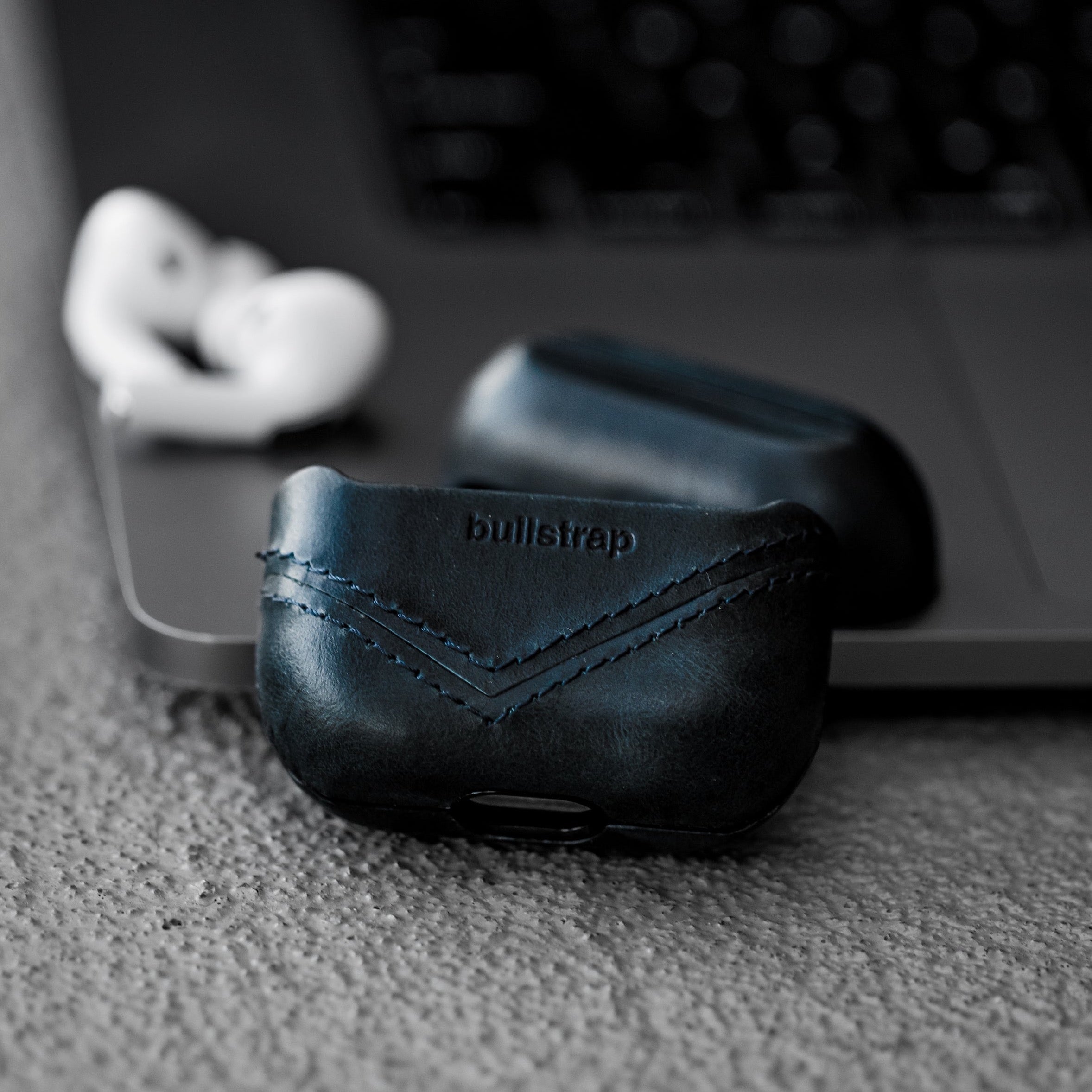Black leather AirPods case with 'bullstrap' logo on a textured surface