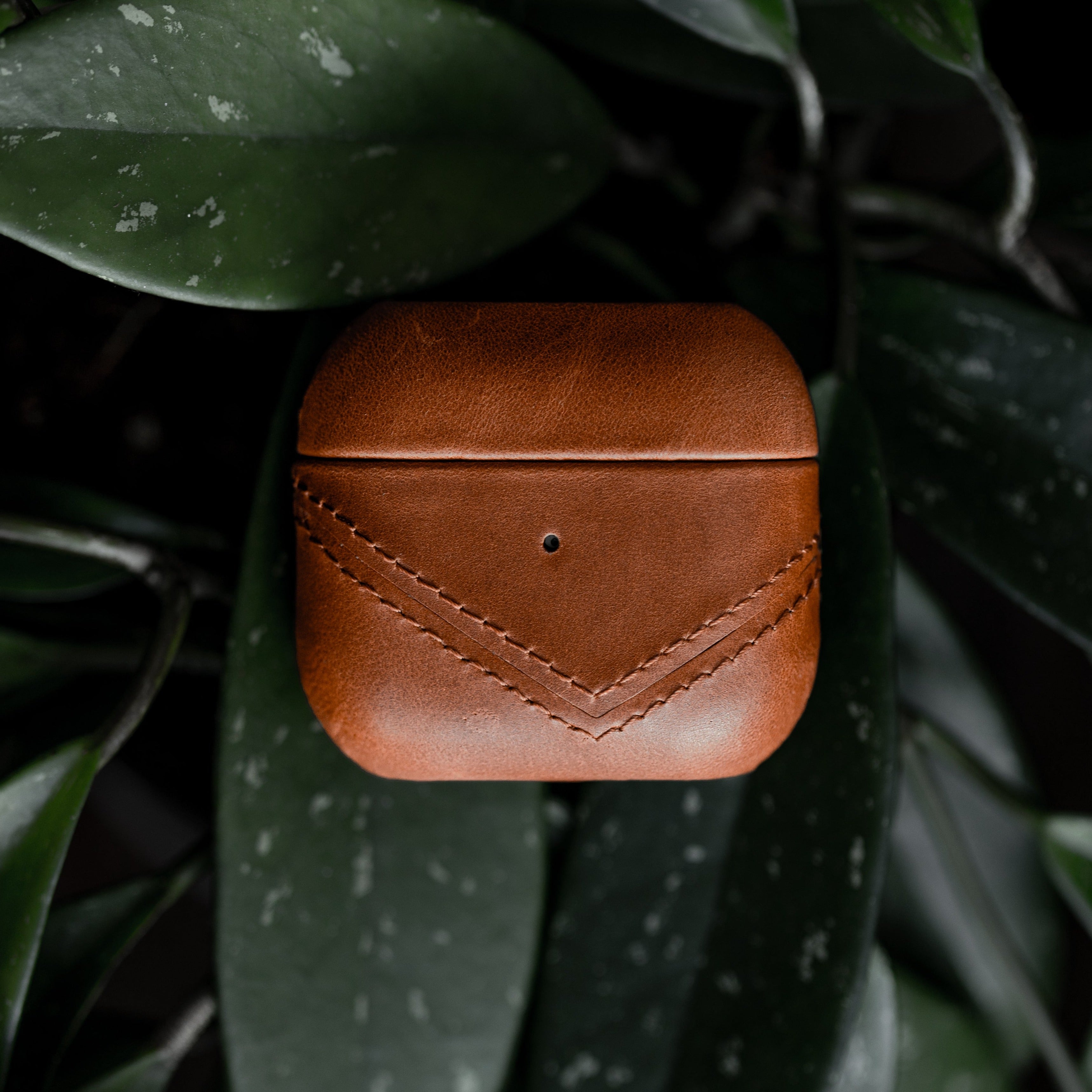 Brown leather AirPods case with a stitched design on a green leaf background.