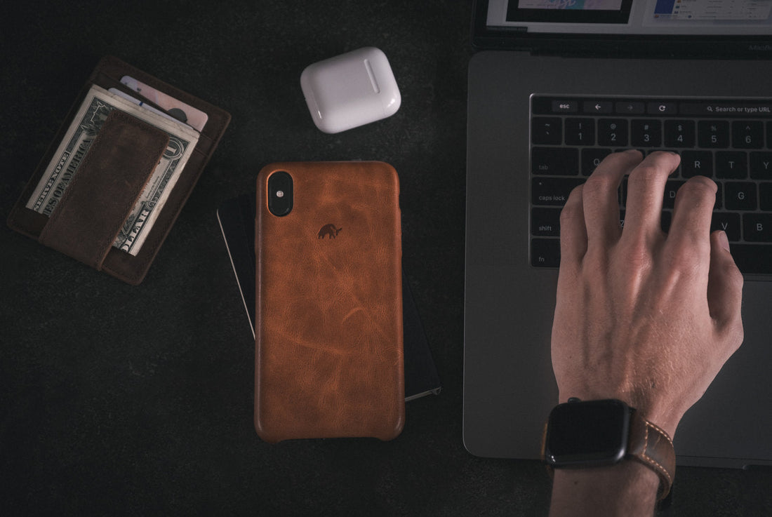A premium leather iPhone case on a table with a wallet, cash, and Apple AirPods, next to a laptop.