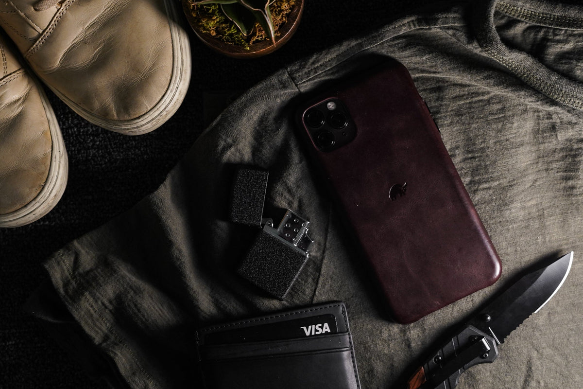 Flat lay of everyday carry: brown leather phone case with bull logo, lighter, wallet showing VISA, folding knife, worn shoes