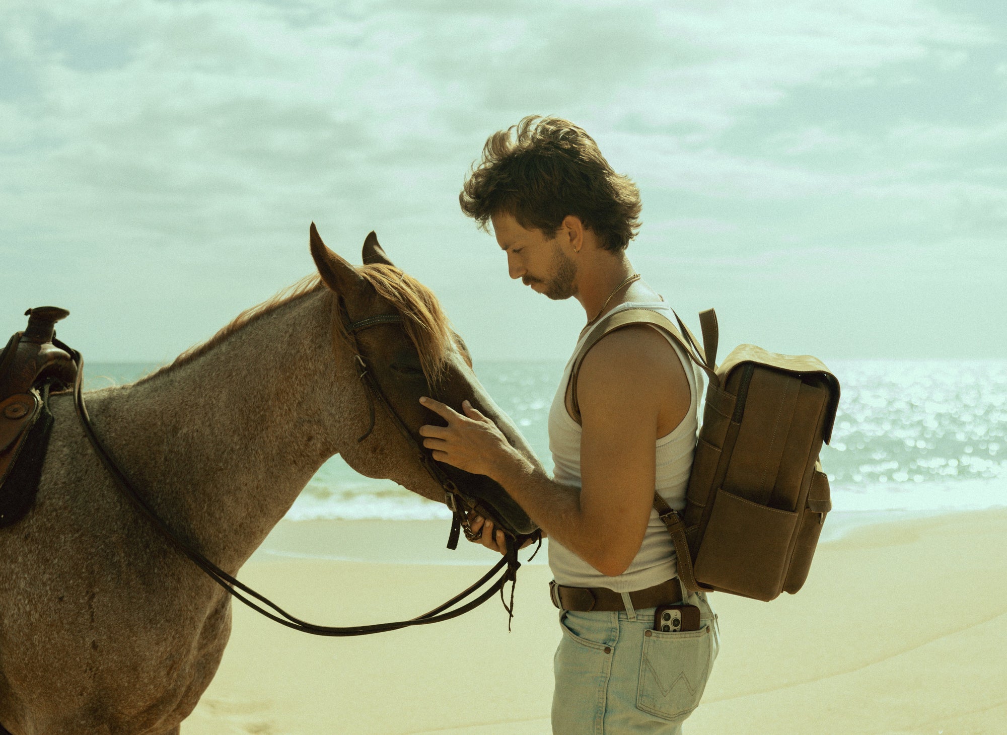 Man wearing a leather backpack gently touching a horse's muzzle on a sunny beach.