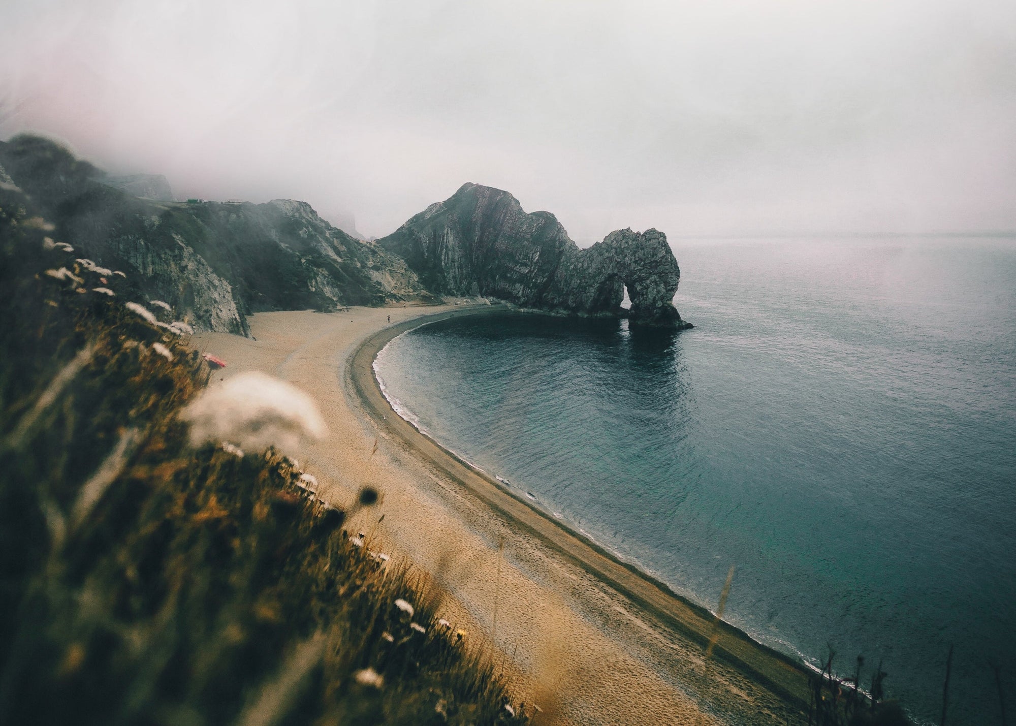 A misty coastal landscape featuring a rocky arch and sandy beach.