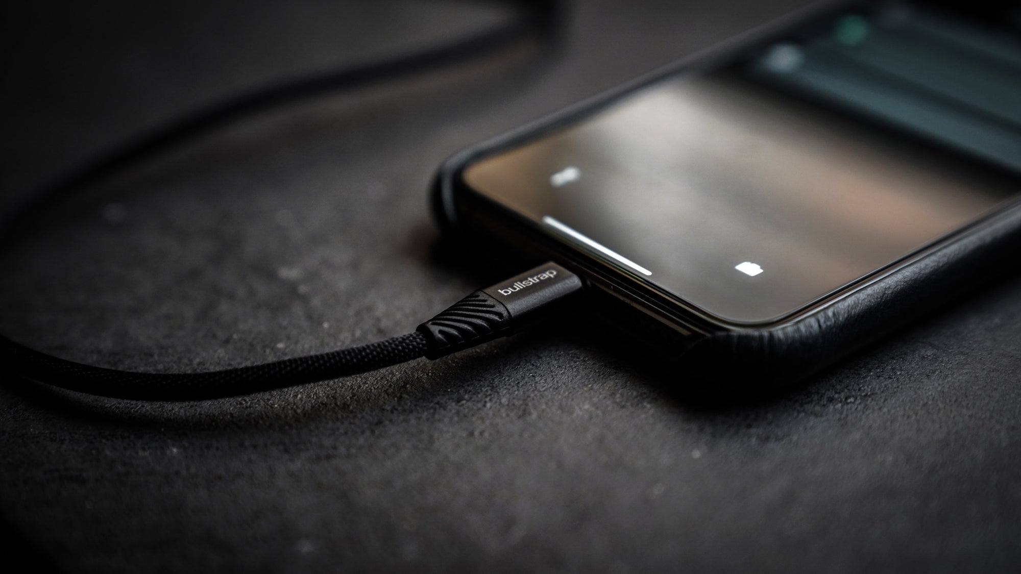 Smartphone connected to a Bullstrap charging cable on a dark surface