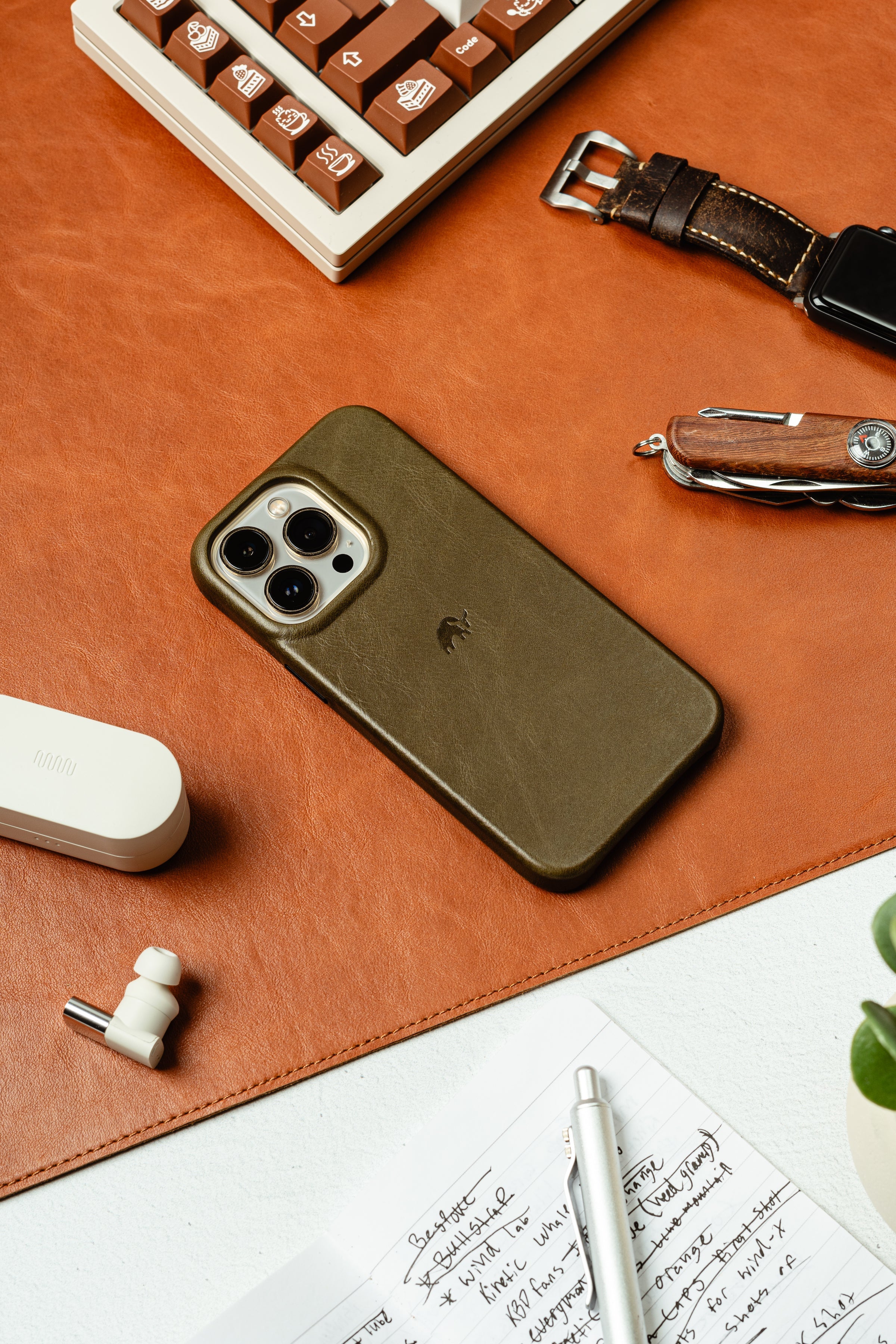 Olive green iPhone case on a desk with a keyboard and notebook