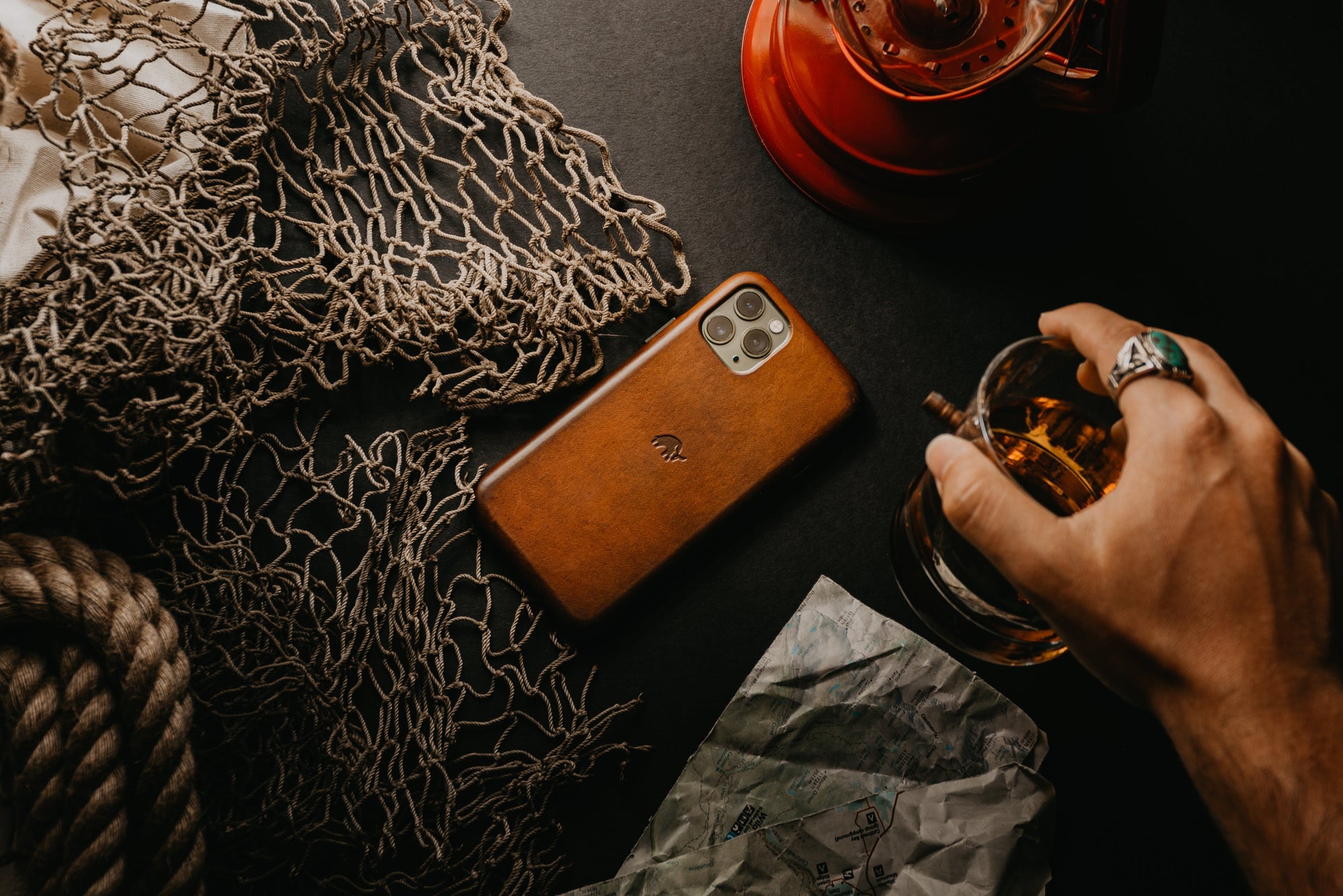 A hand holding a glass next to a phone in a leather case on a dark surface with a lantern and netting.