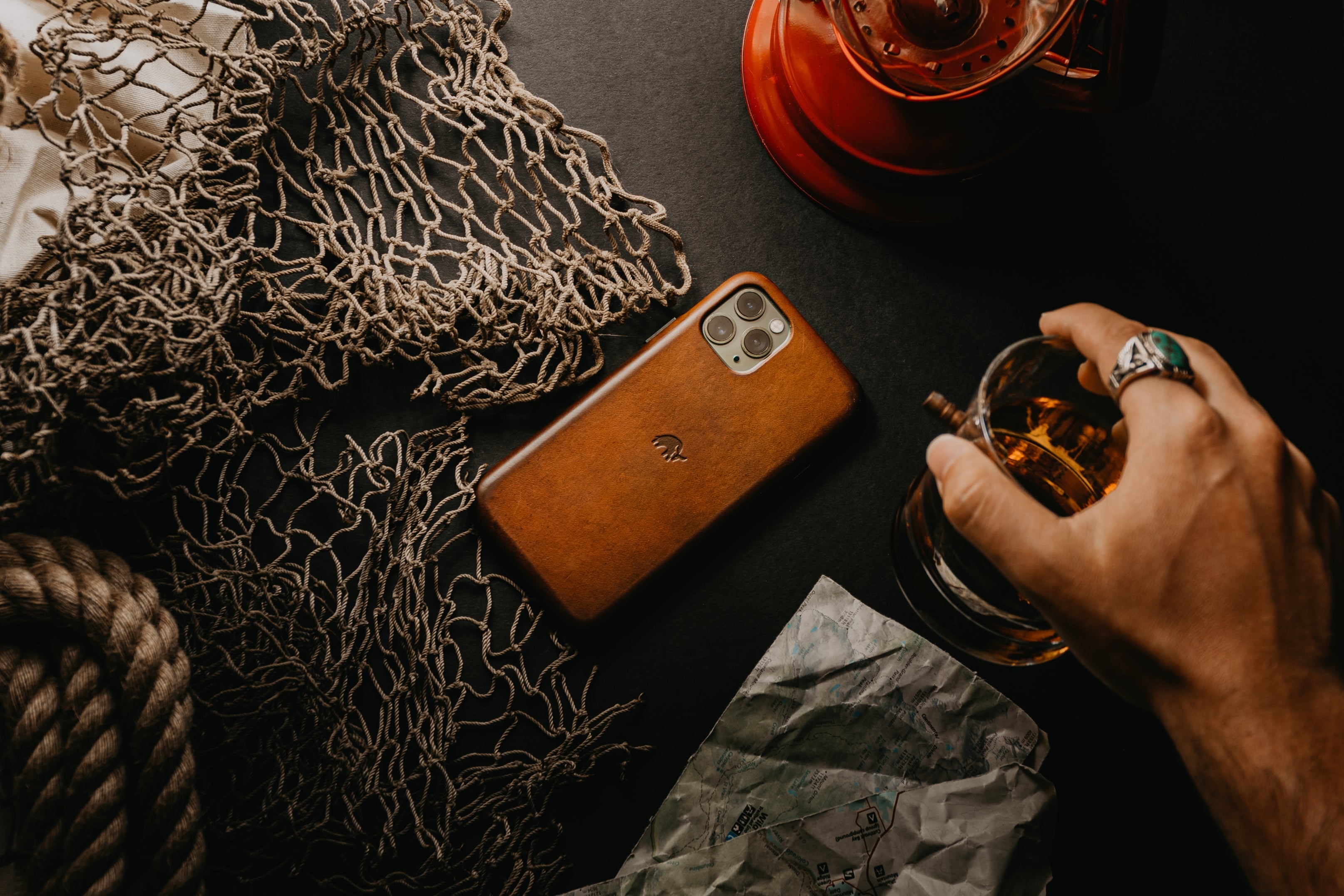 A hand holding a glass next to a phone in a leather case on a dark surface with a lantern and netting.