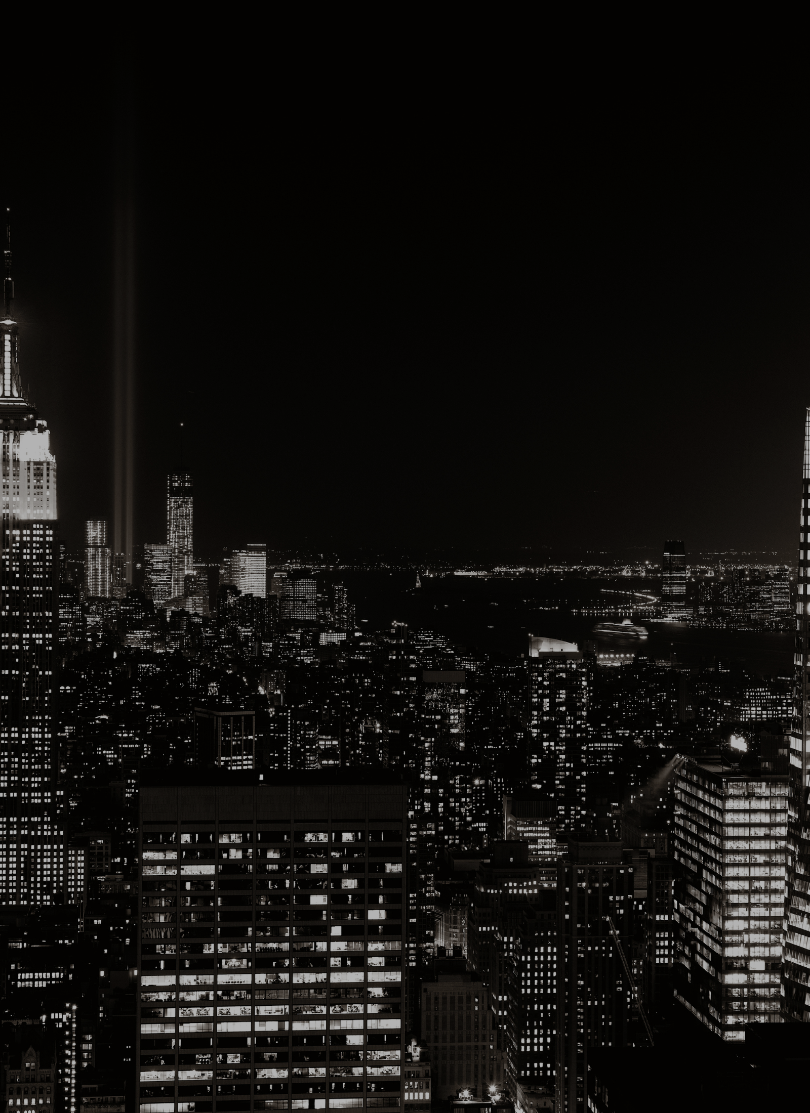 Nighttime cityscape with illuminated skyscrapers and two vertical beams of light in the sky, in black and white.