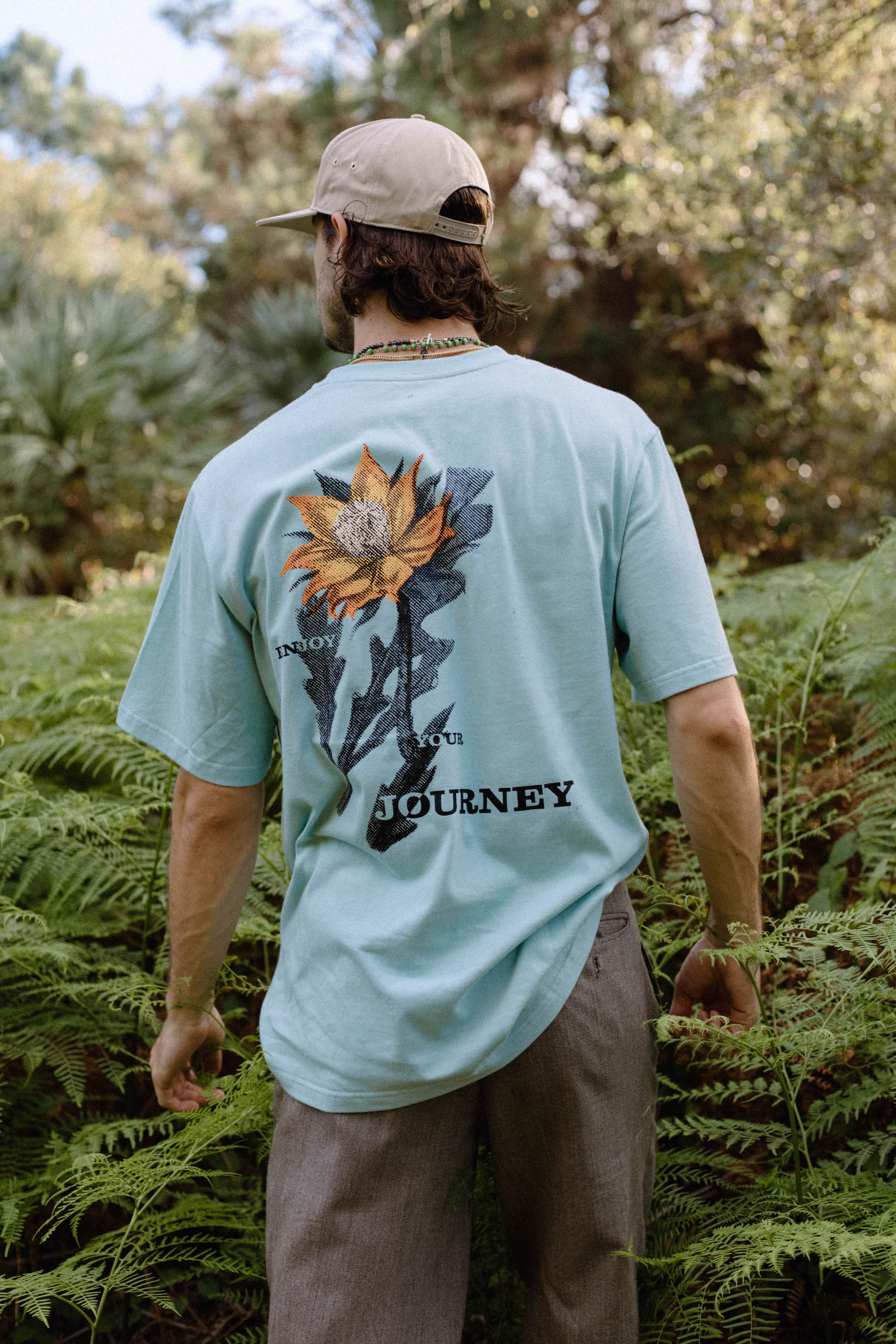 Man wearing a light blue t-shirt with a large sunflower graphic and the words 'ENJOY OUR JOURNEY' on the back, standing in a green forested area.
