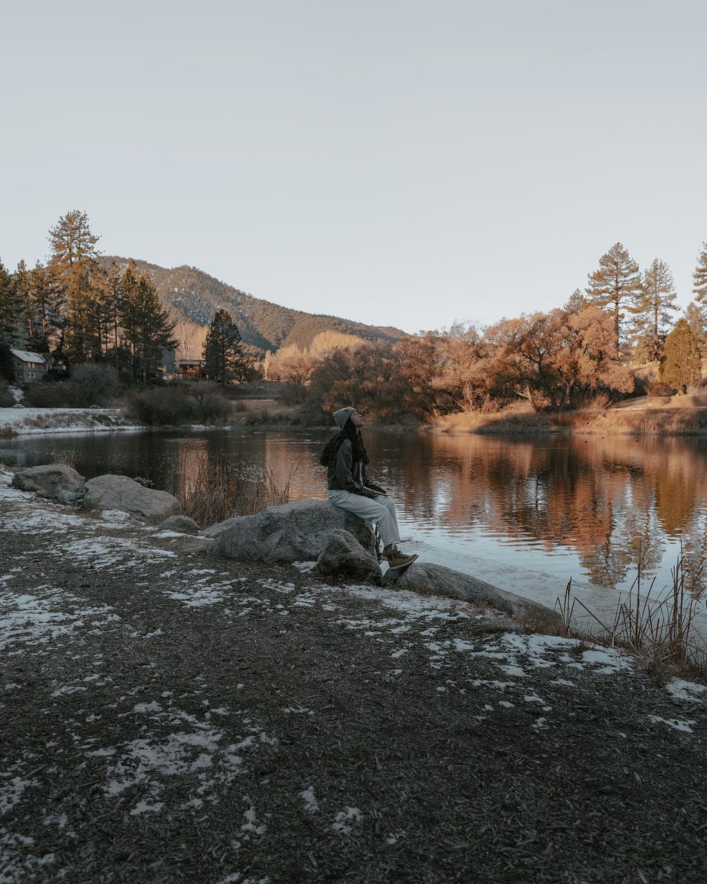 Person sitting on a rock by a lake surrounded by trees and mountains in winter light.
