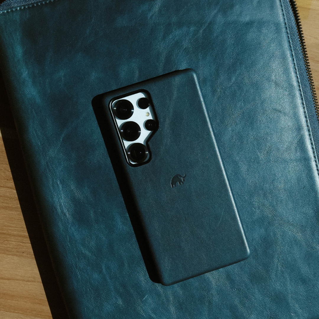 Smartphone in a black leather case showing a four-lens camera array, lying on a teal leather folio with a zipper.