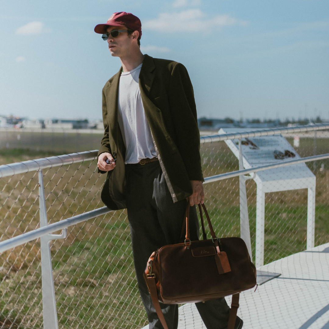 Man wearing red cap and sunglasses, white T-shirt and dark blazer, holding a brown leather duffel bag by a chain-link fence