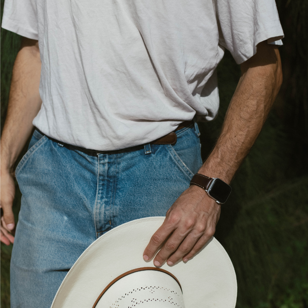 Man wearing a dark brown leather Apple Watch strap on his wrist, holding a white cowboy hat; tucked white tee and blue jeans.