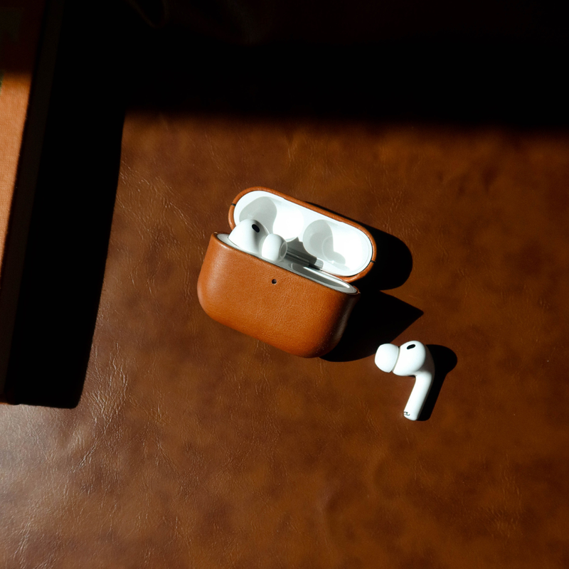 Angled top view of Sienna leather AirPods case with open lid and one AirPod beside it on brown leather background