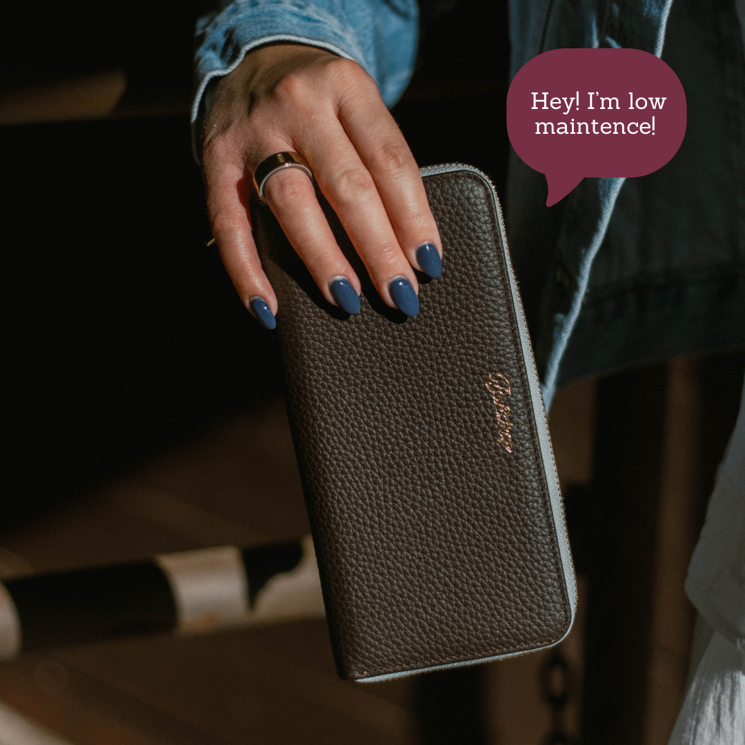 Hand with blue nails holding pebbled zip wallet labeled Bluebelle; speech bubble reads 'Hey! I'm low maintence!'