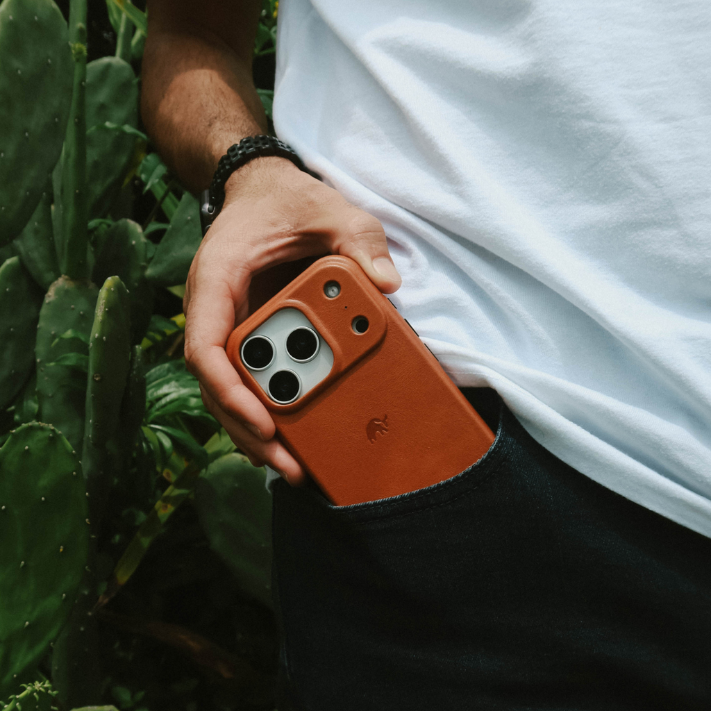 Hand placing phone with sienna leather case into back pocket, close-up showing camera lenses