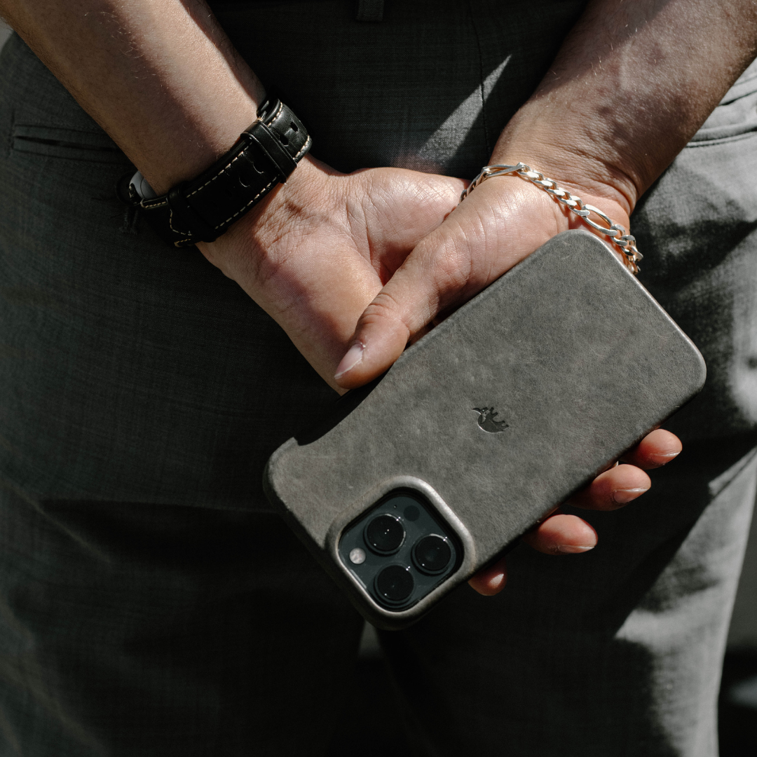 Hands holding smartphone in slate-gray minimalist leather case showing triple camera and small embossed bird logo.