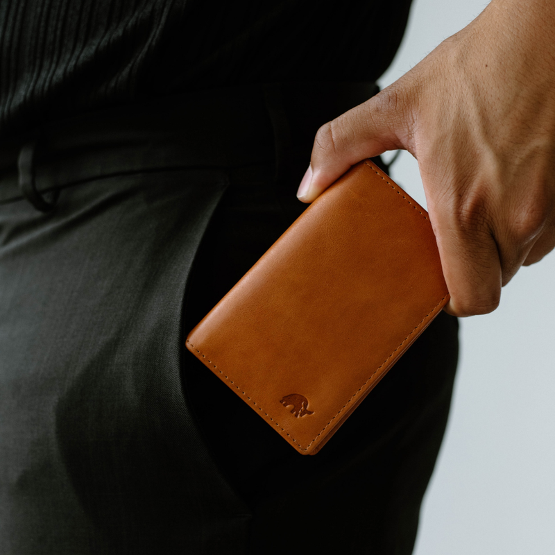 Angled view of Leather Nav Messenger Wallet in sienna showcasing smooth leather, stitched edges, and embossed elephant logo against black pants