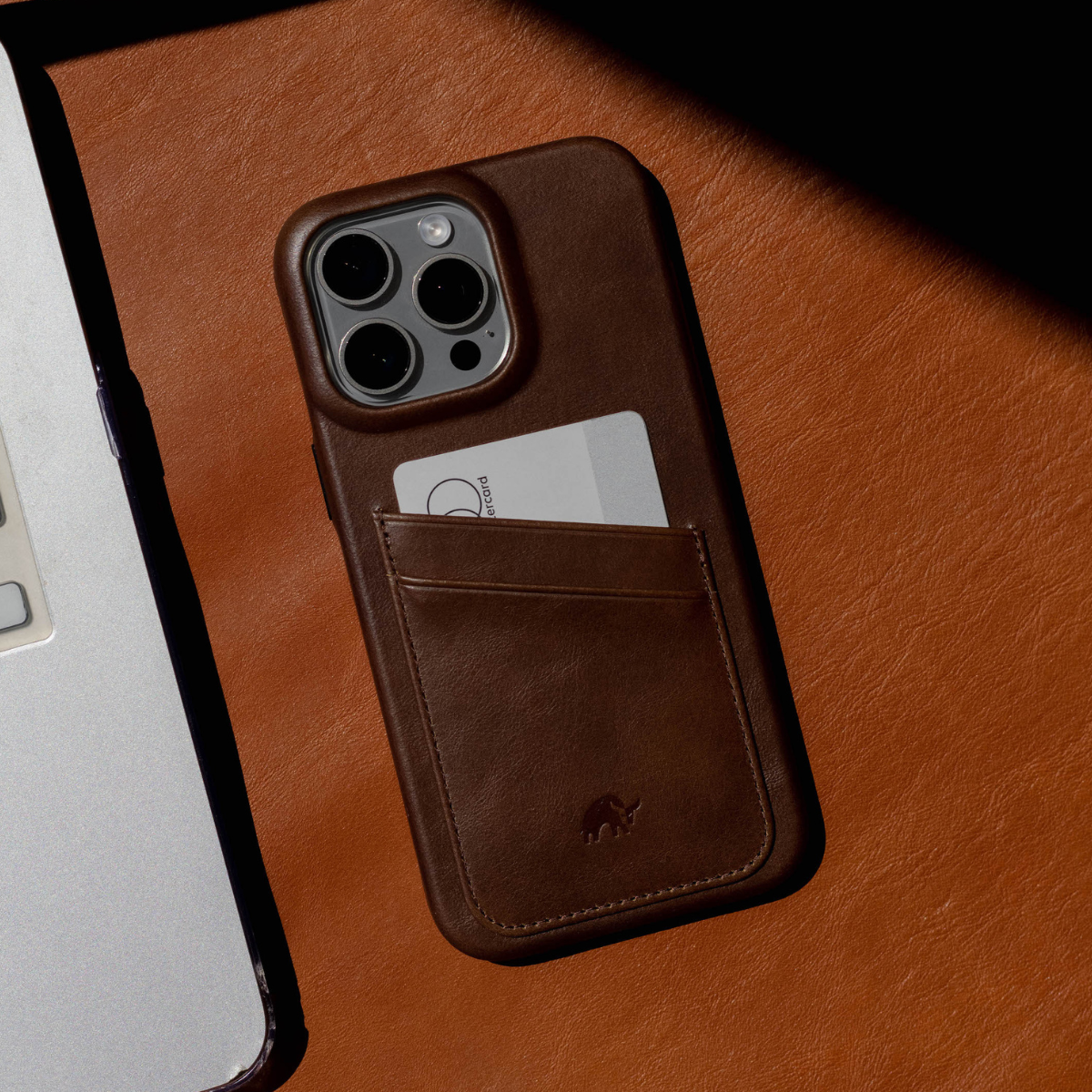 Brown leather portfolio phone case with stitched card pocket holding a card that reads "card", on tan leather surface