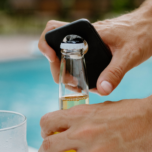 Hand using a black magnetic wallet as a bottle opener to remove a bottle cap beside a pool.