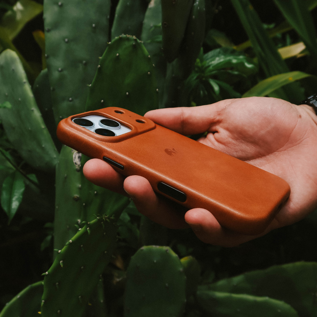 Hand holding sienna leather phone case with triple-camera cutout against green cactus background