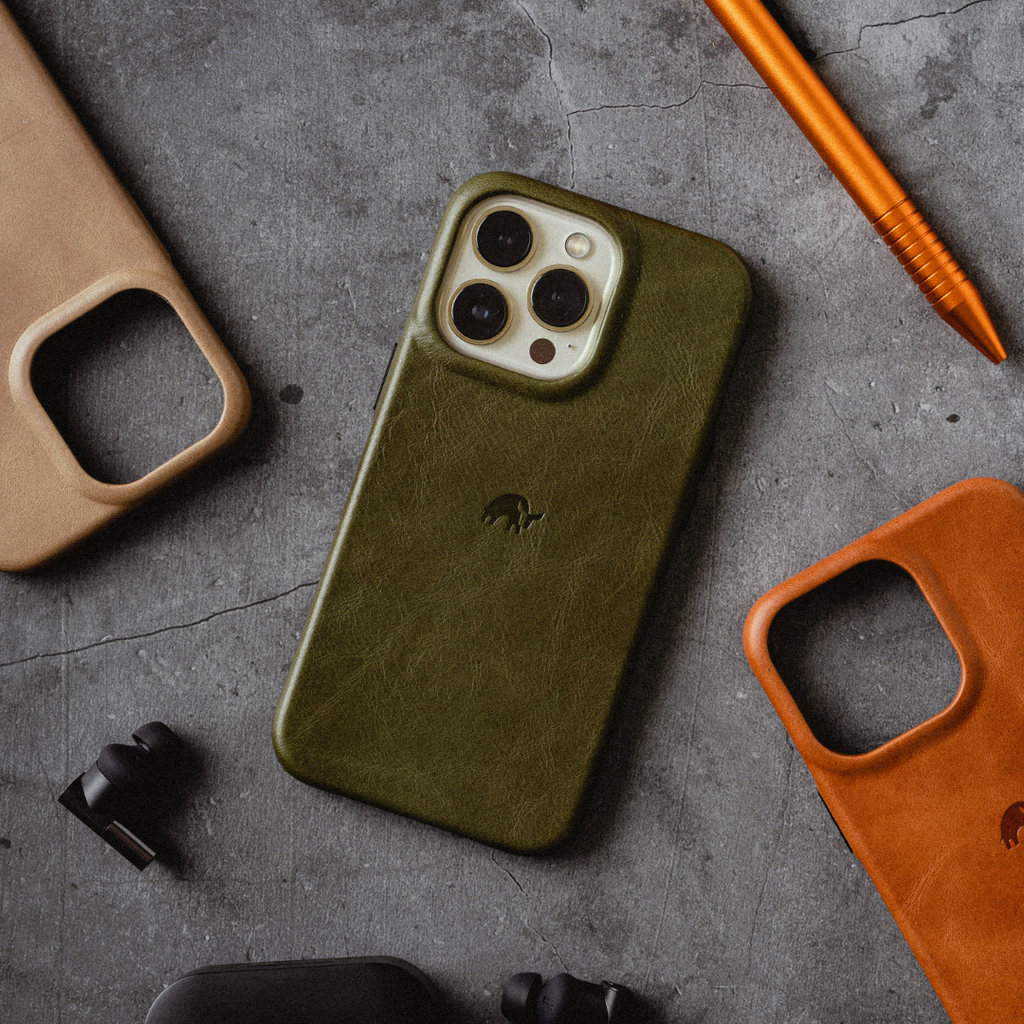 Olive-green leather minimalist phone case (MAVERICK) on textured concrete, surrounded by earbuds and two leather case frames.