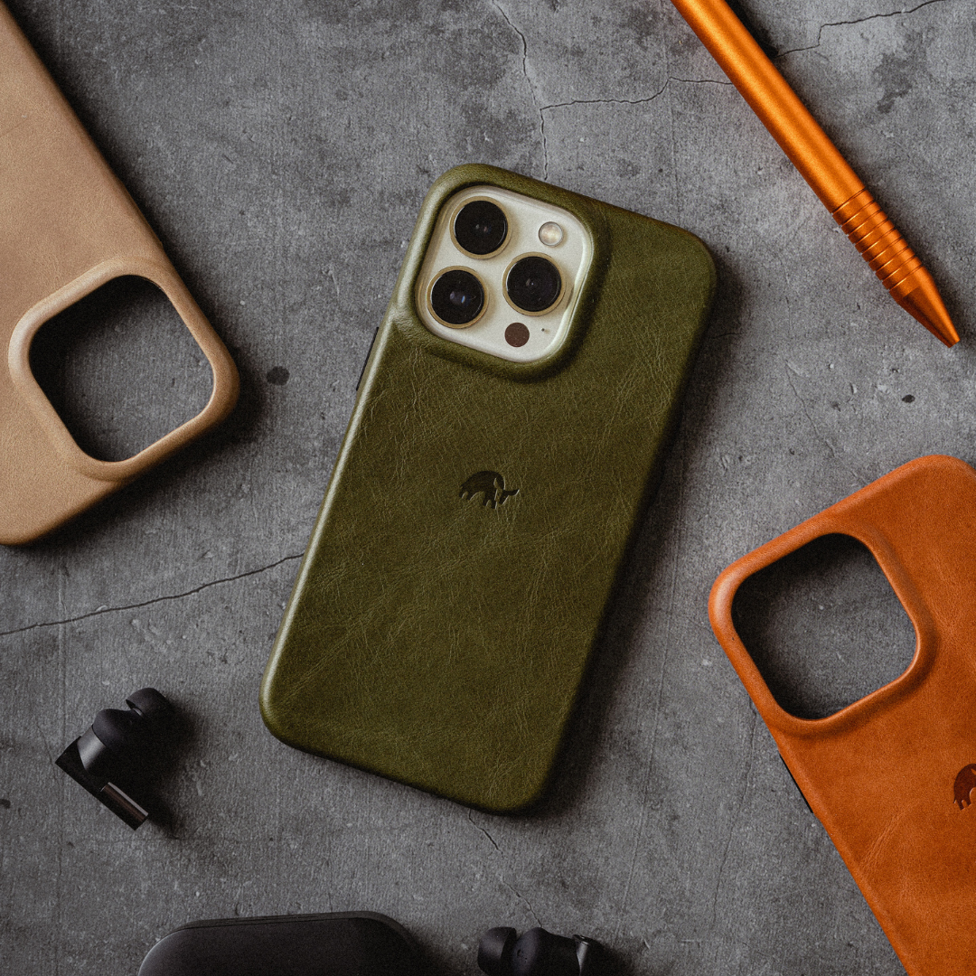 Olive-green leather minimalist phone case (MAVERICK) on textured concrete, surrounded by earbuds and two leather case frames.