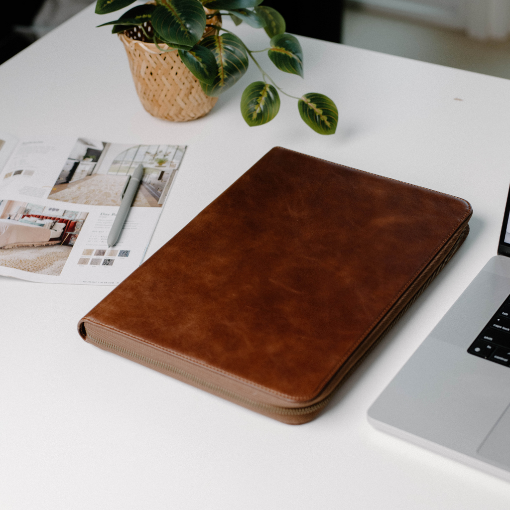 Brown leather MacBook sleeve on a desk next to a laptop and plant
