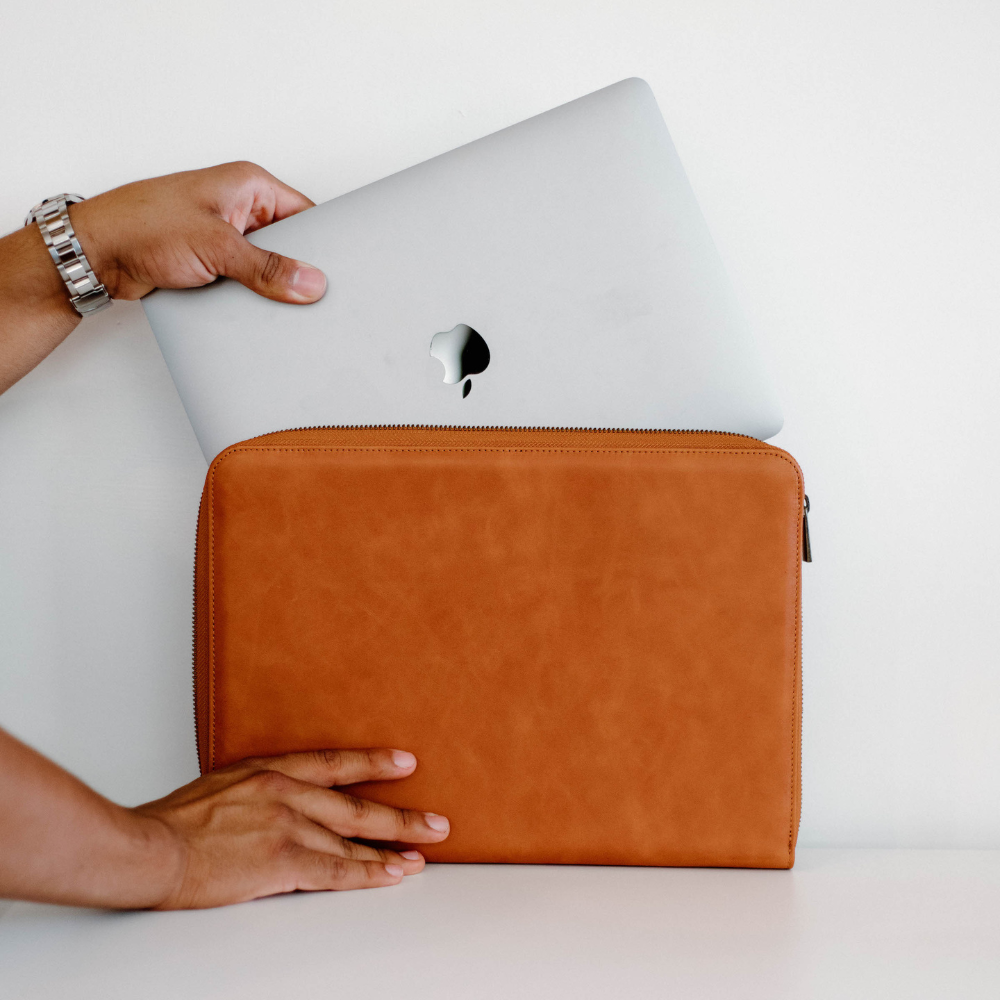 A person holding a tan leather laptop sleeve with a laptop partially inserted
