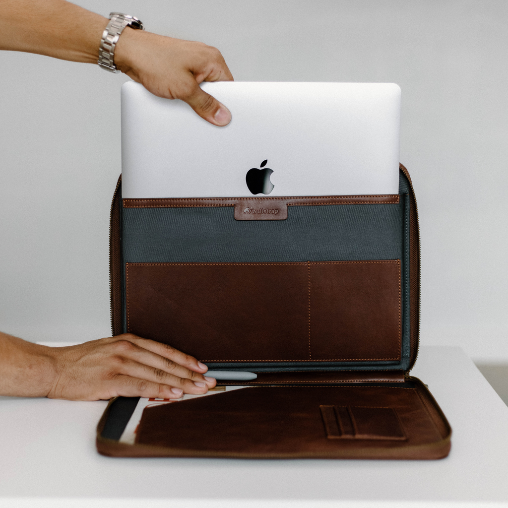 A person holding a MacBook while showing the interior of a leather laptop sleeve with compartments.
