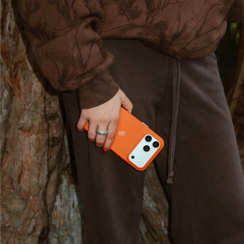 Handheld view of The Performance Case in Blaze showing orange leather and raised camera lip against brown clothing and a tree