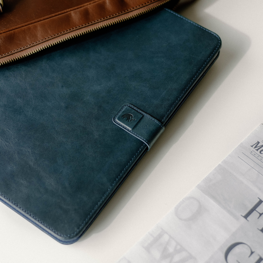 Teal leather iPad folio with snap closure and embossed logo, partially under a brown zippered bag beside a folded newspaper