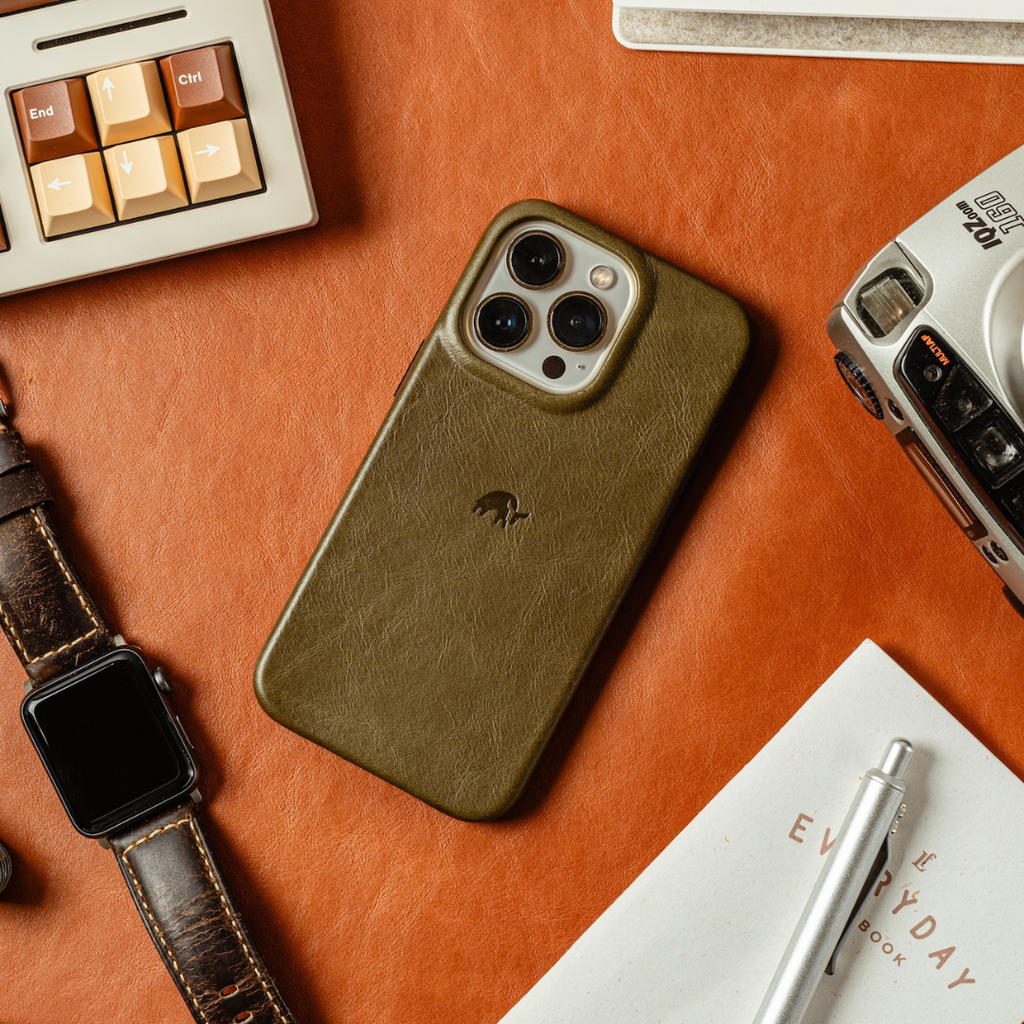 Olive leather iPhone case with embossed logo on orange background beside watch, camera, keyboard; End, Ctrl, EVERYDAY, BOOK
