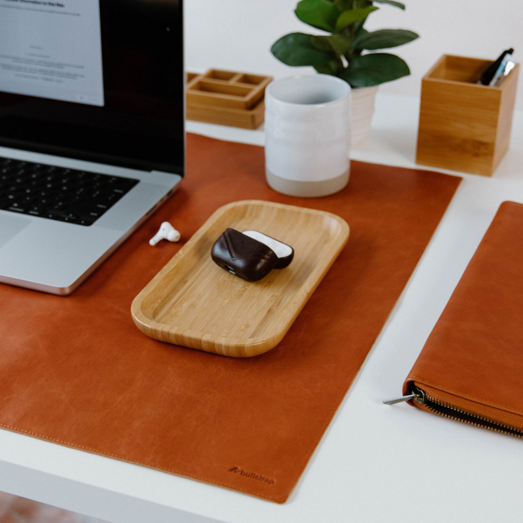 Sienna leather desk mat with laptop, mug, plant, wooden tray holding a dark AirPods case and single earbud.