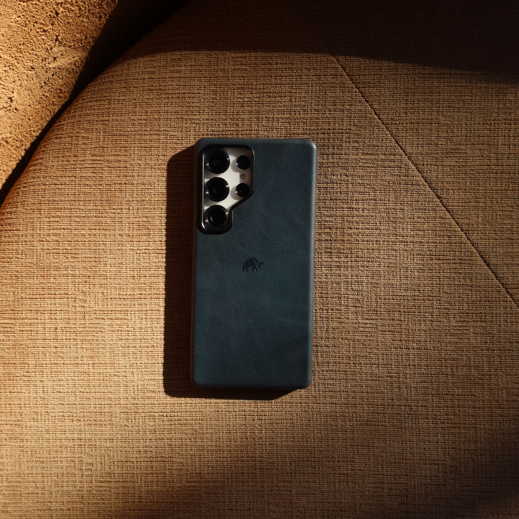 Smartphone with teal leather case and four-lens camera module, embossed Bullstrap logo, resting on brown textured fabric cushion.