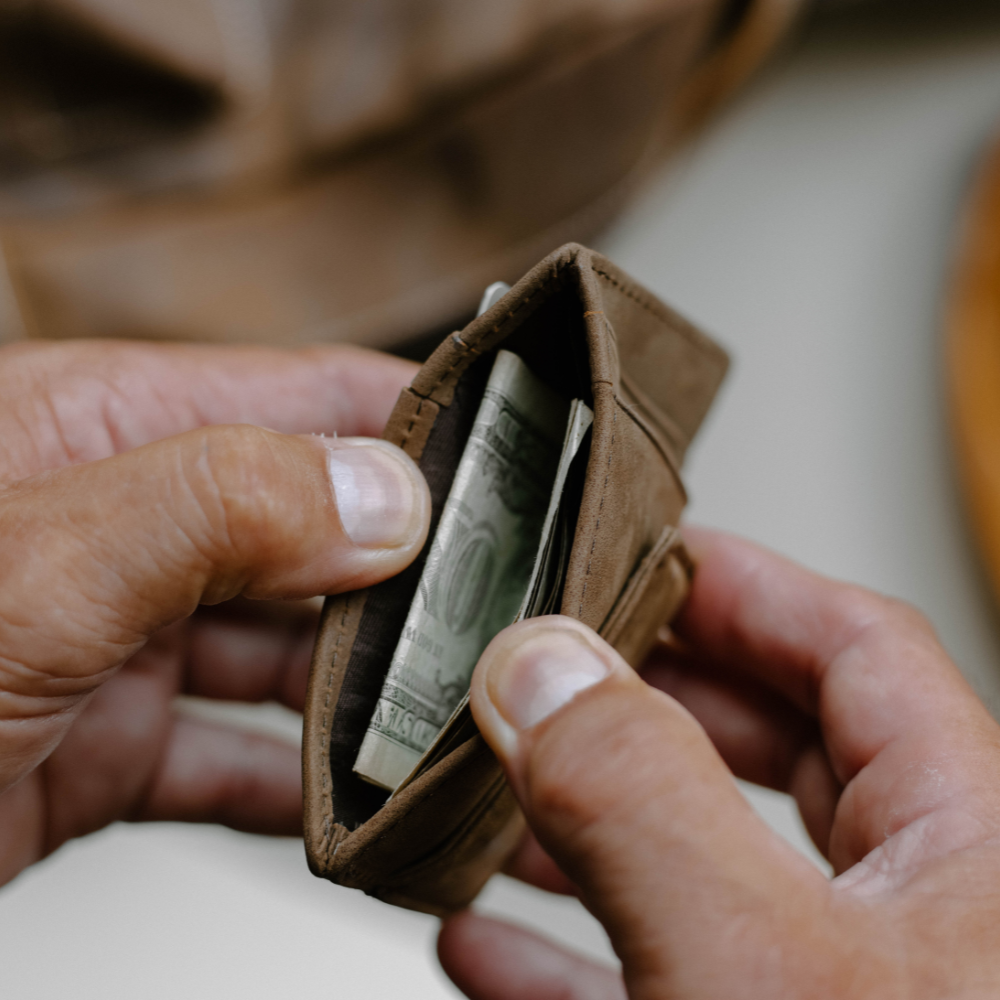 A person holding a brown leather money clip wallet with cash inside.