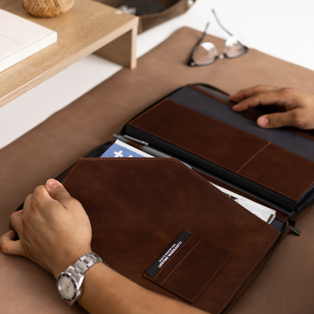 A person holding a brown leather MacBook sleeve open, revealing its interior compartments and documents inside.