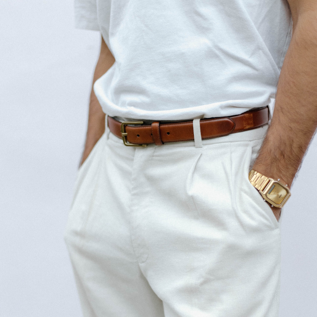Cognac leather belt with brass buckle worn at waist over white trousers, styled with tucked white tee and gold wristwatch