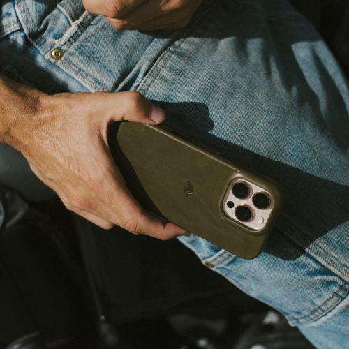 Hand holding iPhone with olive-green leather Maverick minimalist case over denim jeans, triple camera lenses visible