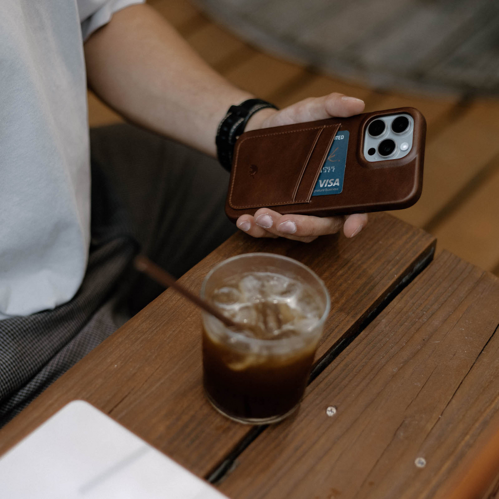 Handheld view of Terra minimalist portfolio case showing rear card pocket and brown leather on a wooden table with iced coffee