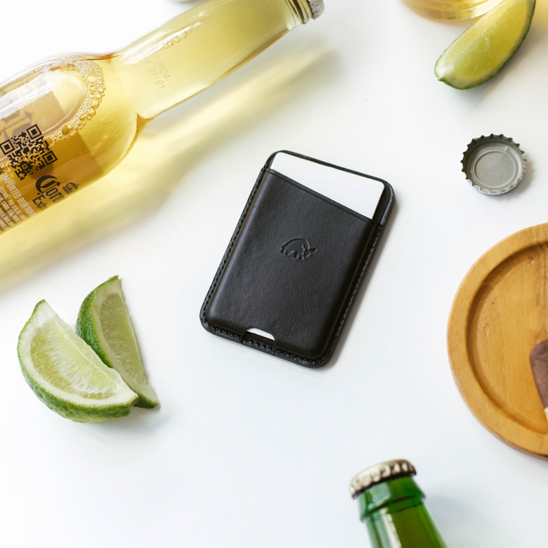 Black leather card wallet holding a white card, beside a Corona Extra bottle, lime wedges and a bottle cap on a white surface.