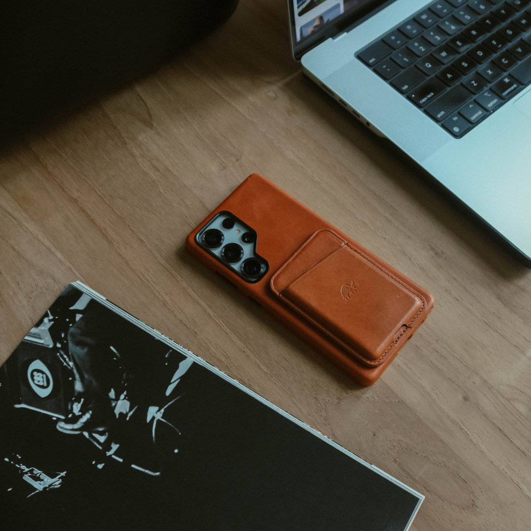 Brown leather smartphone case with built-in card pocket on a wooden desk next to a laptop and a black-and-white magazine.