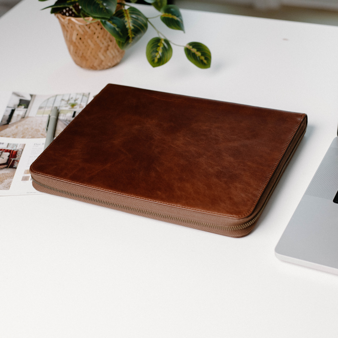Brown leather MacBook sleeve on a white table with a plant and documents nearby.