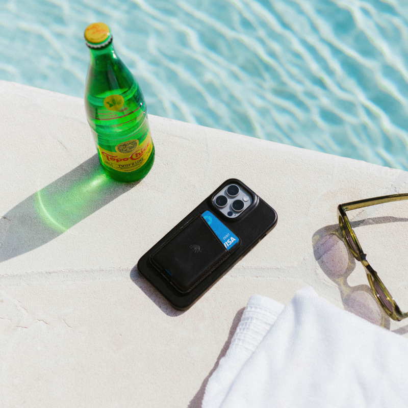 Black phone wallet showing a blue VISA DEBIT card, sunglasses and a Topo Chico bottle labeled TWIST OF LIME beside a pool